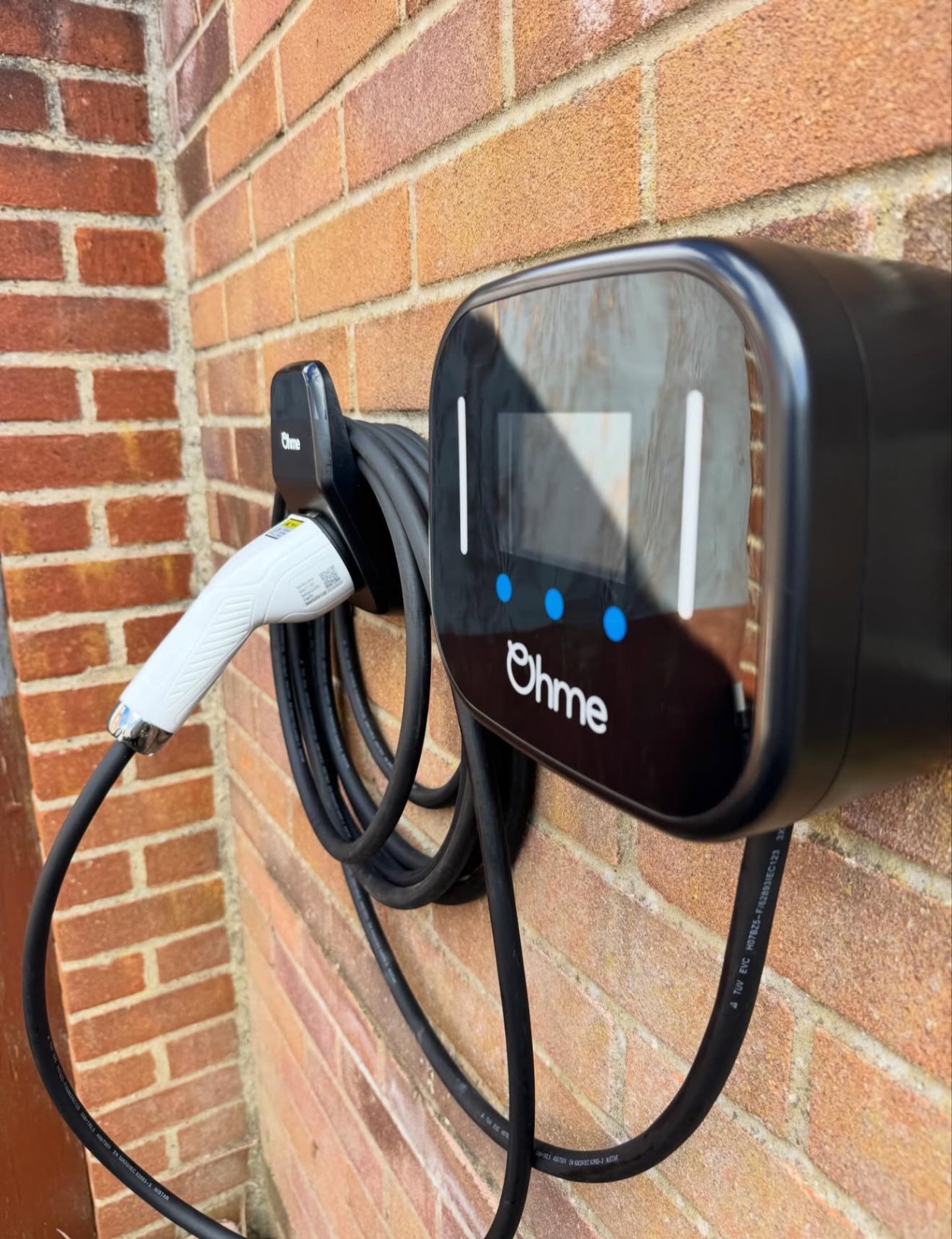 Ohme EV charger with coiled cable and cable holder on a brick wall